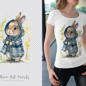 May include: A white t-shirt with a watercolor illustration of a brown rabbit wearing a blue and white plaid raincoat. The rabbit is standing in a field of green grass with yellow flowers. The background is a light blue sky with white raindrops.