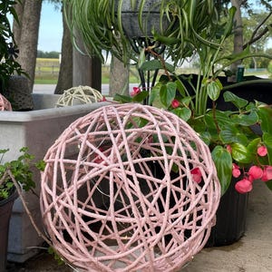 May include: A pink woven sphere with string lights, resting on a concrete base. The decorative orb is surrounded by potted plants, including a spider plant and a begonia with pink flowers. The sphere is a unique home decor item.