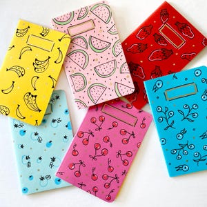 May include: Six small notebooks with different colorful patterns. The notebooks have a gold spine and a gold title bar. The patterns include watermelons, bananas, cherries, blueberries, and strawberries.