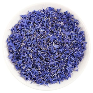 Op de afbeelding: Een witte kom gevuld met levendige blauwe korenbloemblaadjes. De bloemblaadjes zijn een diepe blauwtint met delicate, getextureerde randen. De afbeelding is een bovenaanzicht, dat de ingewikkelde details van de bloemblaadjes laat zien.