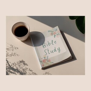 May include: A Bible Study journal with floral accents and the words "Bible Study" in script. A cup of coffee sits next to the journal on a light-colored surface, with shadows from nearby foliage.