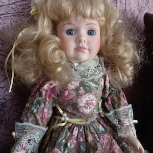 May include: A porcelain doll with blonde, curly hair and blue eyes. The doll wears a floral print dress with lace trim and yellow ribbon accents. The dress features a brown background with pink and white flowers.