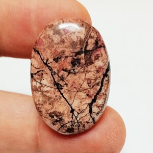 Puede incluir: Una piedra lisa de forma ovalada pulida con un patrón de mármol marrón, rosa y negro. La piedra tiene un acabado brillante.