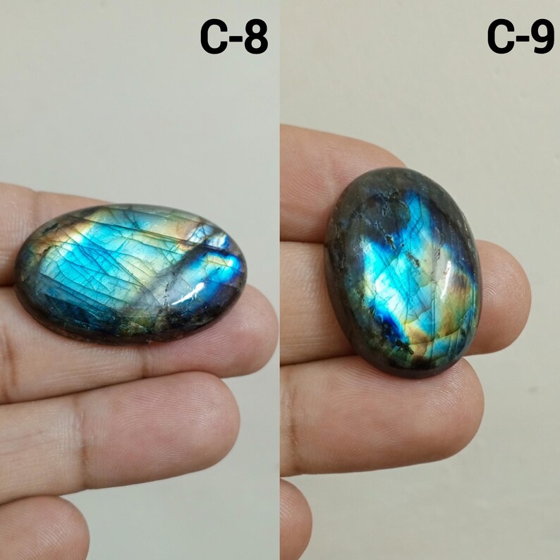 Peut inclure: Deux pierres pr&eacute;cieuses de labradorite de forme ovale. Les pierres pr&eacute;sentent un jeu de couleurs vibrant, avec des &eacute;clairs iris&eacute;s de bleu, de vert et d'or. Les pierres sont tenues dans une main, et le texte "C-8" et "C-9" est visible.