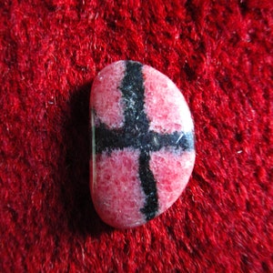 Puede incluir: Una piedra preciosa rosa y negra con un patr&oacute;n de cruz. La piedra preciosa es lisa y pulida.