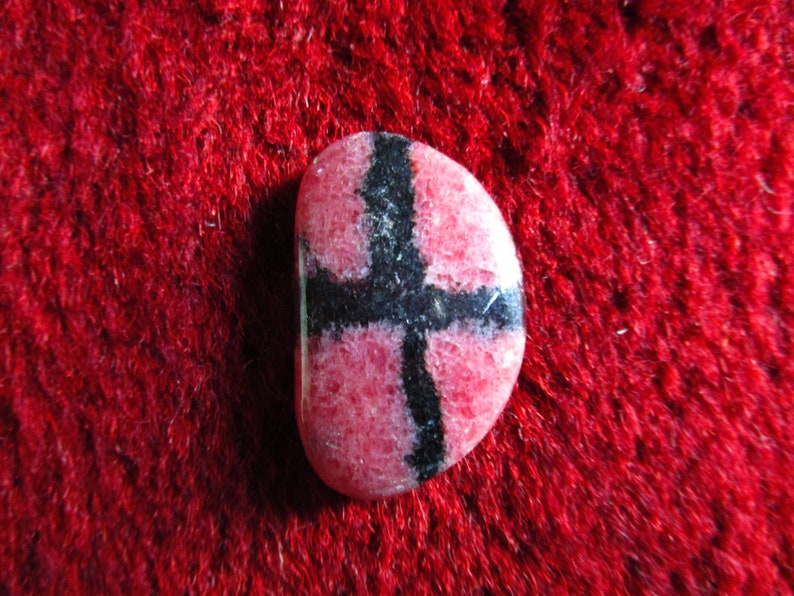 Puede incluir: Una piedra preciosa rosa y negra con un patr&oacute;n de cruz. La piedra preciosa es lisa y pulida.