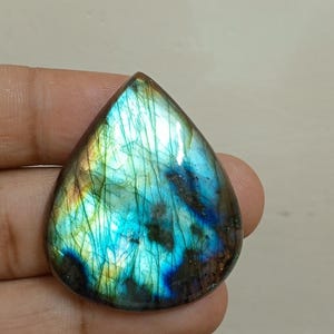Peut inclure: Une pierre pr&eacute;cieuse de labradorite en forme de goutte d'eau, avec des reflets iris&eacute;s bleus, verts et dor&eacute;s. La pierre polie pr&eacute;sente une gamme de couleurs et de motifs internes. La pierre est tenue dans une main.