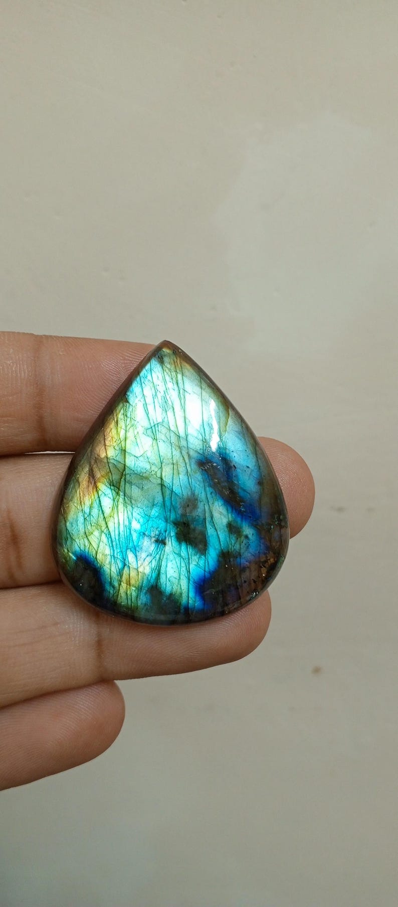 Peut inclure: Une pierre pr&eacute;cieuse de labradorite en forme de goutte d'eau, avec des reflets iris&eacute;s bleus, verts et dor&eacute;s. La pierre polie pr&eacute;sente une gamme de couleurs et de motifs internes. La pierre est tenue dans une main.