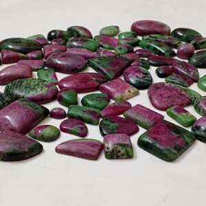 May include: A collection of polished ruby zoisite gemstones in various shapes and sizes. The stones display a mix of deep red and green hues, with some having a marbled appearance. The gemstones are arranged on a white surface.