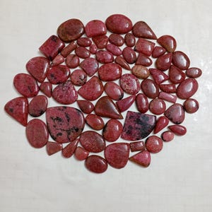 Puede incluir: Una colección de piedras preciosas pulidas de color rojo, de diversas formas y tamaños. Las piedras tienen un acabado brillante y exhiben una gama de tonos, desde el carmesí intenso hasta tonos rosados más claros. Algunas piedras tienen inclusiones oscuras.