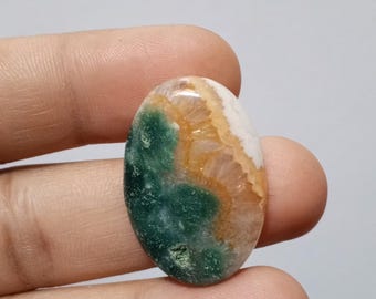 May include: A green, orange and white oval cabochon gemstone with a unique pattern. The gemstone is held in a hand.
