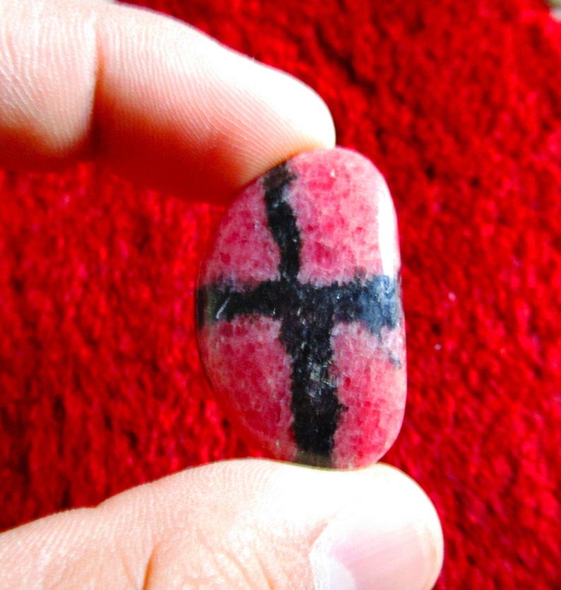Puede incluir: Una piedra preciosa rosa y negra con un patr&oacute;n de cruz. La piedra es lisa y pulida.