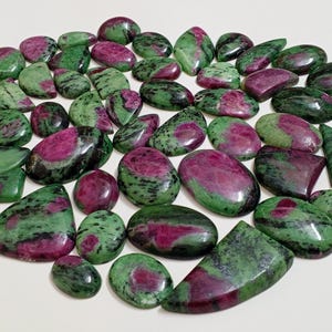 Puede incluir: Una colección de cabujones de piedras preciosas verdes y rosas. Las piedras son de varias formas y tamaños, y tienen un aspecto moteado.