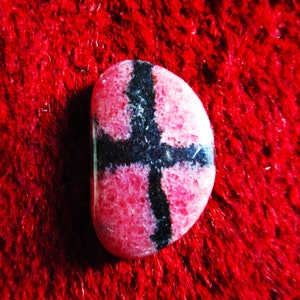 Puede incluir: Una piedra preciosa rosa y negra con un patr&oacute;n de cruz. La piedra preciosa es lisa y pulida.