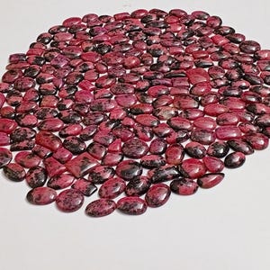 Puede incluir: Una colección de piedras preciosas pulidas en tonos rojo oscuro y negro. Las piedras varían en forma, incluyendo formas ovaladas, rectangulares e irregulares. Las piedras preciosas están dispuestas muy juntas, creando una superficie texturizada.