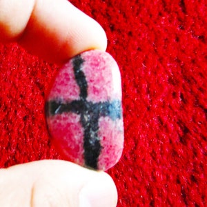 Puede incluir: Una piedra preciosa rosa y negra con un patr&oacute;n de cruz. La piedra es lisa y pulida.