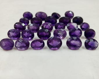 Könnte beinhalten: Eine Ansammlung oval geschliffener Amethyst-Edelsteine. Die facettierten Steine zeigen eine tiefviolette Farbe mit Variationen in Farbe und Klarheit. Die Edelsteine sind auf einer weißen Oberfläche angeordnet.