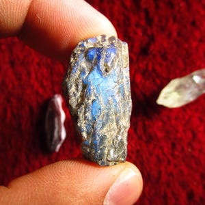 May include: A rough-cut, blue and gray labradorite gemstone is held between fingers, showcasing its iridescent flash. Two other gemstones, one white and one purple, are also visible against a red background. The labradorite is the focal point.