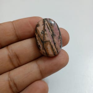 Puede incluir: Una gema ovalada con una superficie pulida. La piedra presenta una mezcla de colores rosa, marrón y negro, con un patrón de rayas y vetas. La gema se sostiene en una mano, mostrando su tamaño y forma.