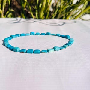 Puede incluir: Un collar de cuentas turquesas. El collar está compuesto por pequeñas cuentas de turquesa de corte cuadrado. Las cuentas son de un tono azul vibrante y están ensartadas para formar una forma circular. El fondo es una superficie blanca con follaje verde.