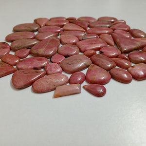 Puede incluir: Colección de piedras preciosas de rodonita rosa pulidas de varias formas y tamaños. Las piedras muestran una gama de tonos rosas con vetas más oscuras, dispuestas sobre una superficie blanca. Las piedras son lisas y brillantes.