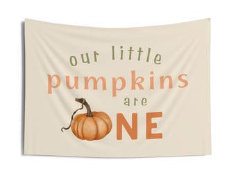 May include: A cream-colored wall hanging with the text "our little pumpkins are ONE" in green and orange lettering. A watercolor-style orange pumpkin is featured below the text, perfect for fall decor.