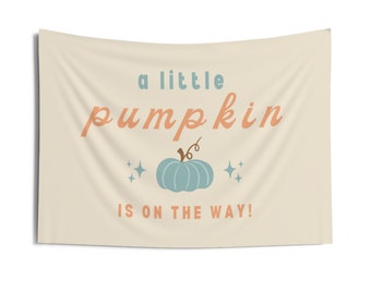 May include: A beige banner with the text "a little pumpkin is on the way!" in orange. A blue pumpkin with a green stem is in the center of the banner with two small white stars on either side.