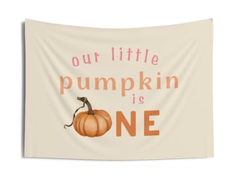 Può includere: Uno striscione beige con il testo "our little pumpkin is ONE" in rosa e arancione. Una piccola zucca arancione con un gambo verde è al centro dello striscione.