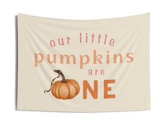 May include: A beige banner with the text "our little pumpkins are one" in pink and orange. A small orange pumpkin with a green stem is in the center of the banner.