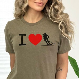 以下が含まれることがあります： 「I love」という文字と赤いハートが書かれた緑色のTシャツ。ハートの右側には、フィールドホッケー選手のシルエットがあります。
