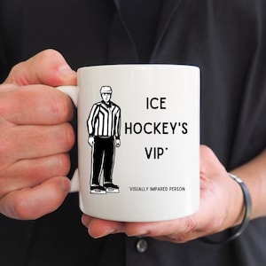 May include: White ceramic coffee mug with a black and white illustration of an ice hockey referee. The text on the mug reads "ICE HOCKEY'S VIP*" with the asterisk next to VIP followed by the text "VISUALLY IMPAIRED PERSON".