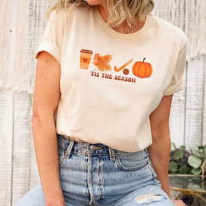 Peut inclure: Un t-shirt beige avec un motif graphique repr&eacute;sentant une tasse de caf&eacute;, une feuille, un b&acirc;ton de hockey et une citrouille. Le texte "'Tis the season" est imprim&eacute; sous le graphique.