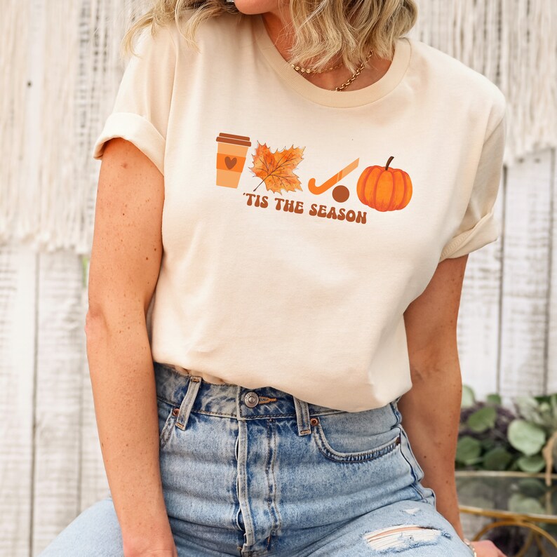 Peut inclure: Un t-shirt beige avec un motif graphique repr&eacute;sentant une tasse de caf&eacute;, une feuille, un b&acirc;ton de hockey et une citrouille. Le texte "'Tis the season" est imprim&eacute; sous le graphique.