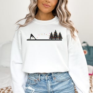 May include: White sweatshirt featuring a black silhouette of a curling player, colorful lights, and three Christmas trees. The design is printed on a crew neck sweatshirt. The sweatshirt is made of a soft material.