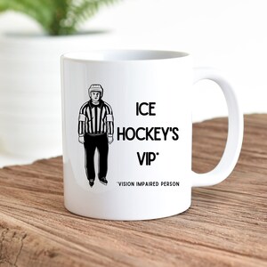 May include: White ceramic coffee mug with a black and white illustration of an ice hockey referee. The text reads "ICE HOCKEY'S VIP*" with the text "VISION IMPAIRED PERSON" below.