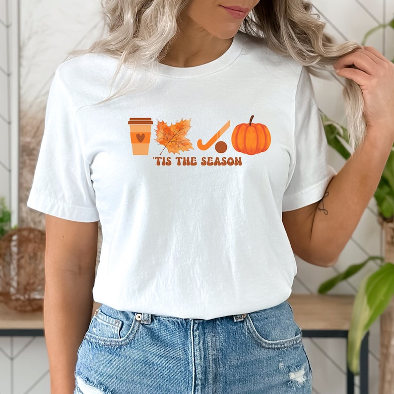 Peut inclure: T-shirt blanc avec un motif repr&eacute;sentant une tasse de caf&eacute;, une feuille, un b&acirc;ton de hockey et une citrouille. Le texte "'Tis the season" est imprim&eacute; sous le motif.