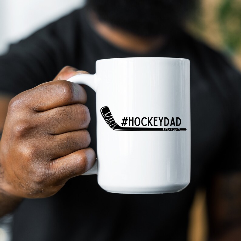 Op de afbeelding: Een witte keramische koffiemok met een zwarte hockeystick en de tekst "#HOCKEYDAD" erop gedrukt.