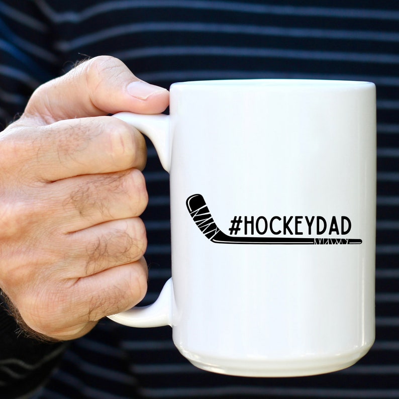 Op de afbeelding: Witte keramische koffiemok met een zwarte hockeystick en de tekst "#HOCKEYDAD" erop gedrukt.