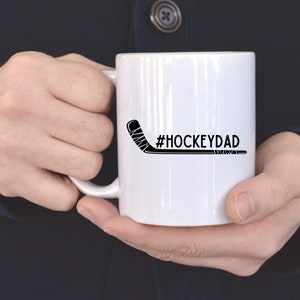 Op de afbeelding: Witte keramische mok met een zwarte hockeystick en de tekst "#HOCKEYDAD" erop gedrukt.