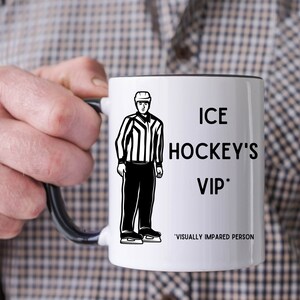 May include: White ceramic mug with a black rim and handle. The mug features a black and white illustration of an ice hockey referee standing on skates. The text "ICE HOCKEY'S VIP*" is printed on the mug, with the asterisk indicating "VISUALLY IMPAIRED PERSON".