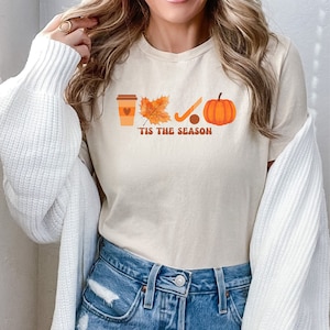 Peut inclure: Un t-shirt beige avec un motif graphique repr&eacute;sentant une tasse de caf&eacute;, une feuille, un b&acirc;ton de hockey, une rondelle et une citrouille. Le texte "'Tis the season" est imprim&eacute; sous le graphique.