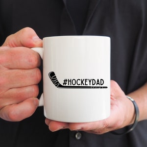 Op de afbeelding: Witte keramische koffiemok met een zwarte hockeystick-afbeelding en de tekst "#HOCKEYDAD".
