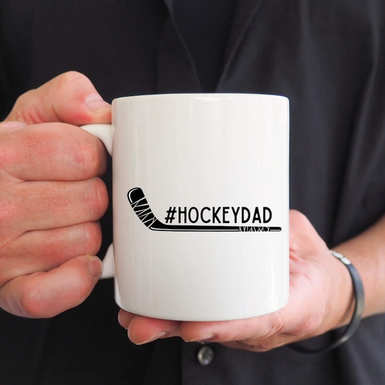 Op de afbeelding: Witte keramische koffiemok met een zwarte hockeystick-afbeelding en de tekst "#HOCKEYDAD".
