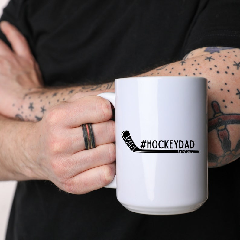 Op de afbeelding: Een witte keramische mok met een zwarte hockeystick en de tekst "#HOCKEYDAD" erop gedrukt.
