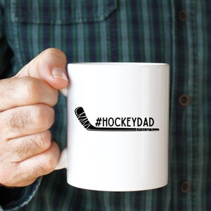 Op de afbeelding: Een witte keramische mok met een zwarte hockeystick en de tekst "#HOCKEYDAD" erop gedrukt.