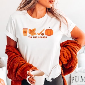 Peut inclure: T-shirt blanc avec un motif graphique repr&eacute;sentant une tasse de caf&eacute;, une feuille, un b&acirc;ton de hockey, une rondelle de hockey et une citrouille. Le texte "Tis the Season" est imprim&eacute; sous le graphique.