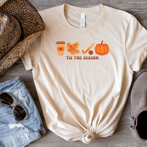 Peut inclure: Un t-shirt beige avec un motif graphique repr&eacute;sentant une tasse de caf&eacute;, une feuille, un b&acirc;ton de hockey et une citrouille. Le texte "'Tis the season" est imprim&eacute; sous le graphique.