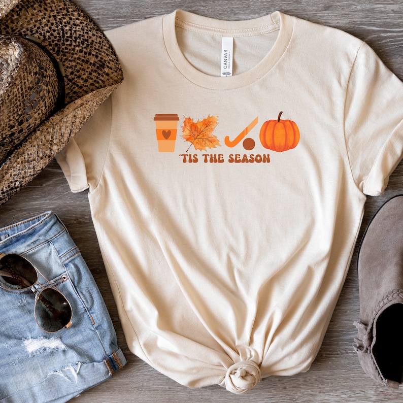 Peut inclure: Un t-shirt beige avec un motif graphique repr&eacute;sentant une tasse de caf&eacute;, une feuille, un b&acirc;ton de hockey et une citrouille. Le texte "'Tis the season" est imprim&eacute; sous le graphique.