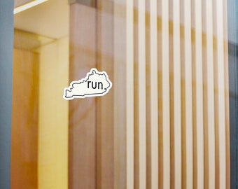 Könnte beinhalten: Ein weißer Aufkleber in Form von Kentucky mit dem Wort "run" in schwarzer Schrift. Der Aufkleber befindet sich auf einer Glasoberfläche, mit einem Hintergrund aus Holzpaneelen und einer gelben Wand.