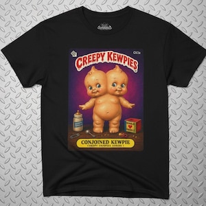 May include: A black t-shirt featuring a graphic print of a two-headed doll. The text reads CREEPY KEWPIES, CONJOINED KEWPIE, and CREEPY KEWPIES SERIES 1. The illustration includes a baby bottle, a small spoon, and a red block with a heart on it.